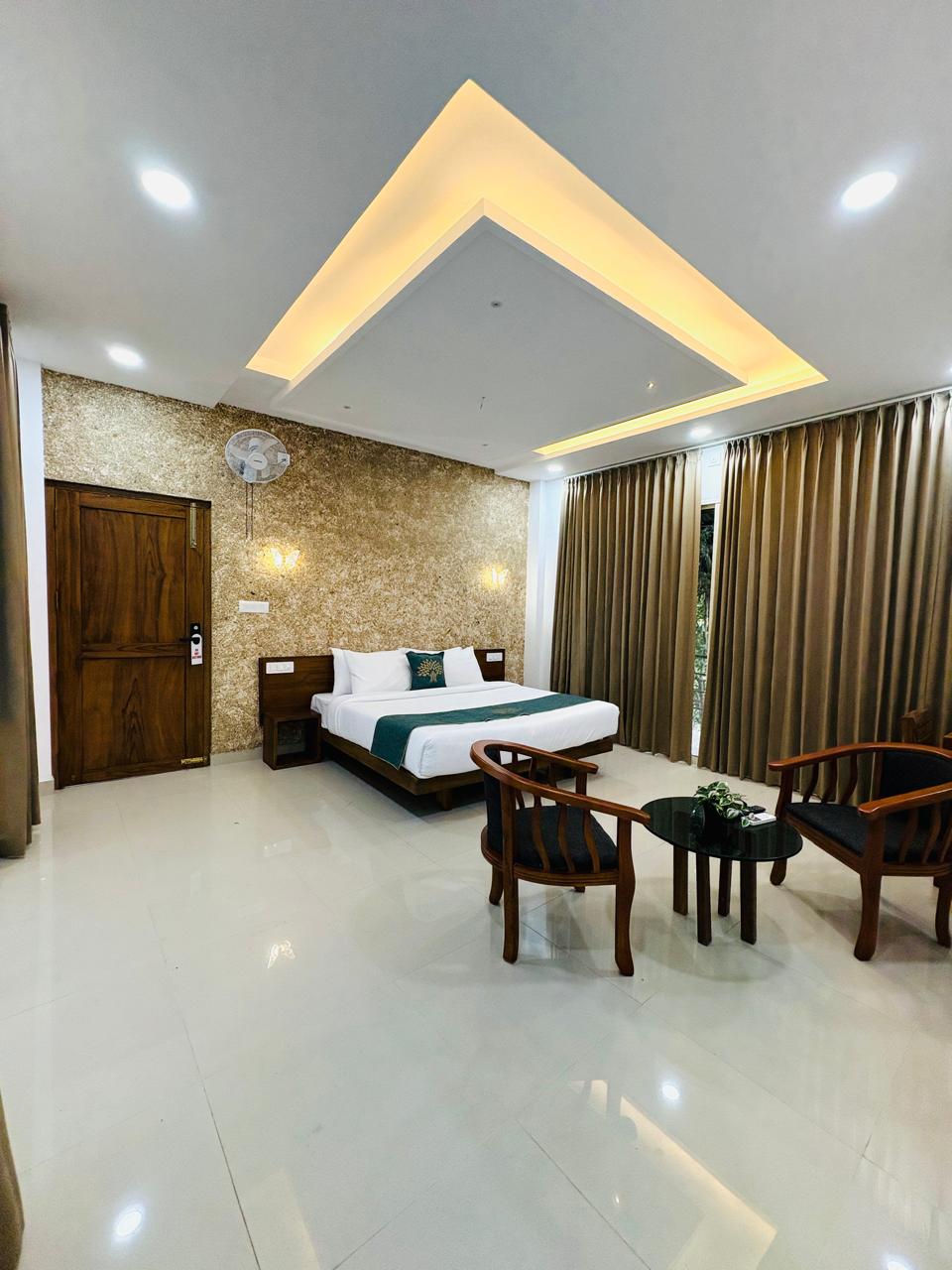 Spacious modern hotel room at JKR Gardens featuring a king-size bed with crisp white linens and a teal runner, wooden furniture, and floor-to-ceiling curtains allowing natural daylight. The room includes a tiled floor, yellow accent wall, air conditioning, and a window overlooking lush green trees, creating a peaceful and inviting atmosphere ideal for relaxation. No visible text.